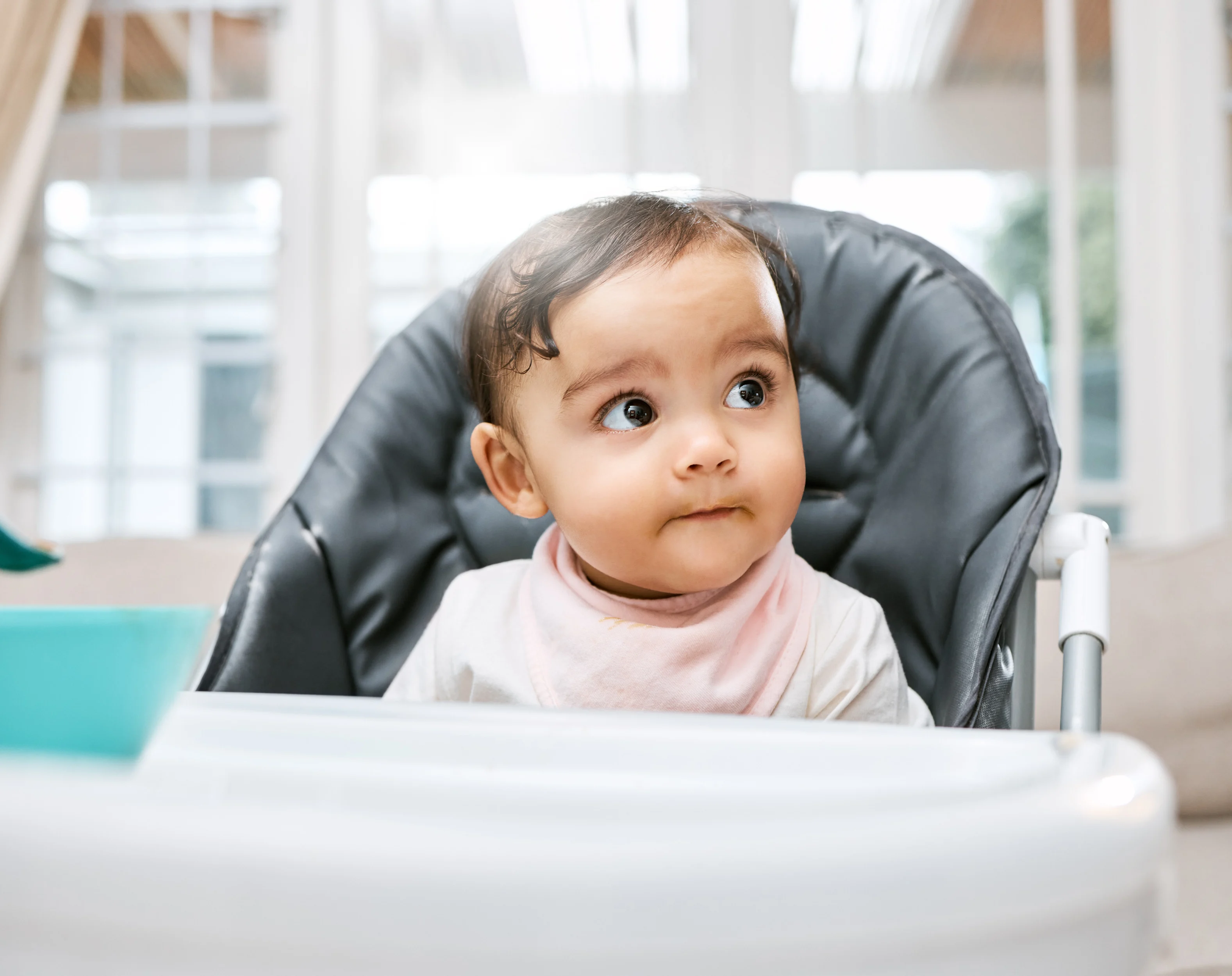 A baby wearing a pink bib sits in a dark grey high chair, looking up curiously, with a bright sunlit room in the background.