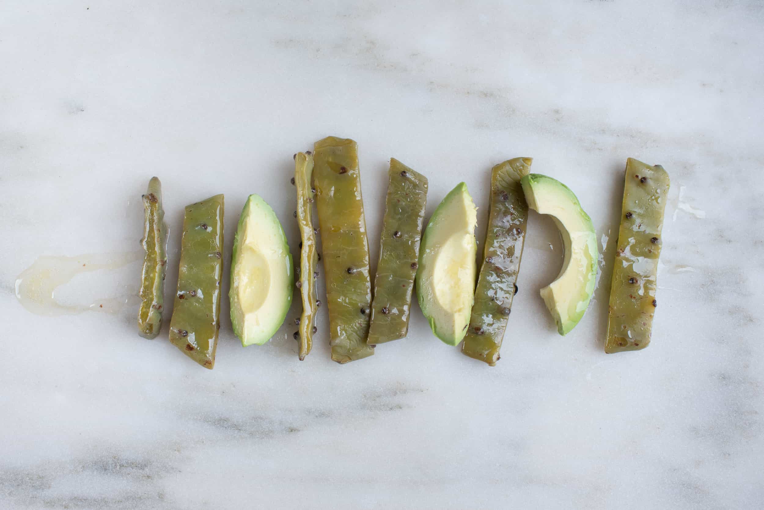 thin rectangular strips of cooked nopales and thin wedges of avocado on a white background drizzled with oil