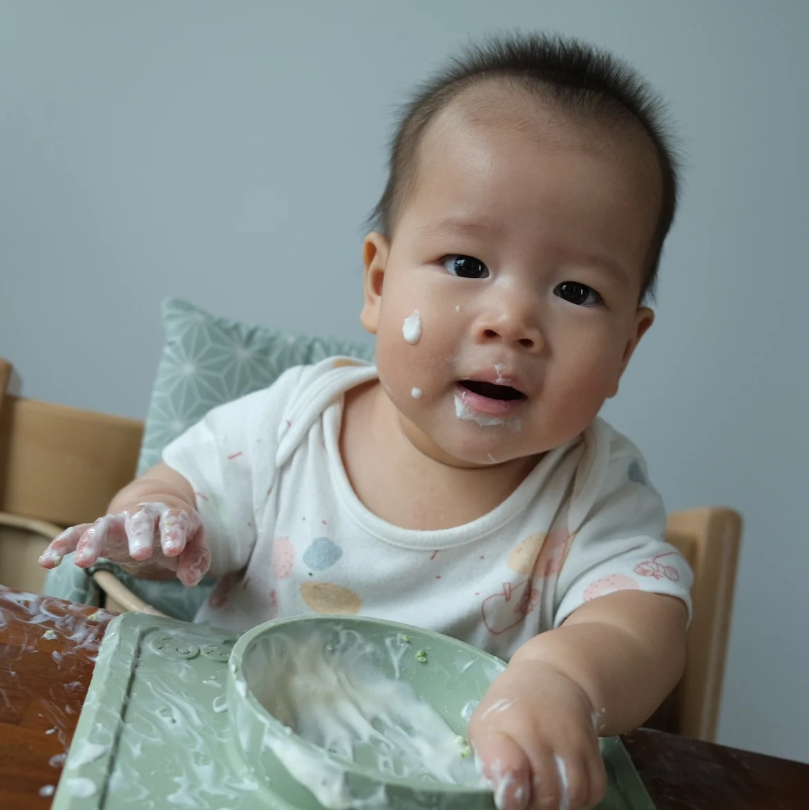 A baby with yogurt on their face and hands sits in a high chair, smiling with curiosity. A green bowl and mat are in front, conveying playful chaos.