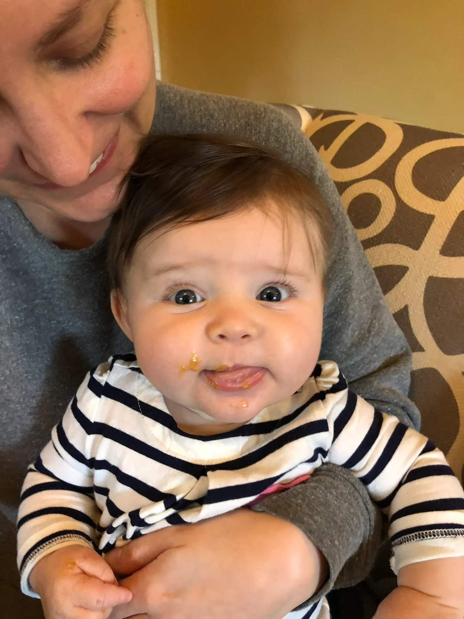 a photograph of a five month old baby on her mother's lap. the baby is licking a bit of peanut butter from the side of her mouth, that she's just been fed by her mother
