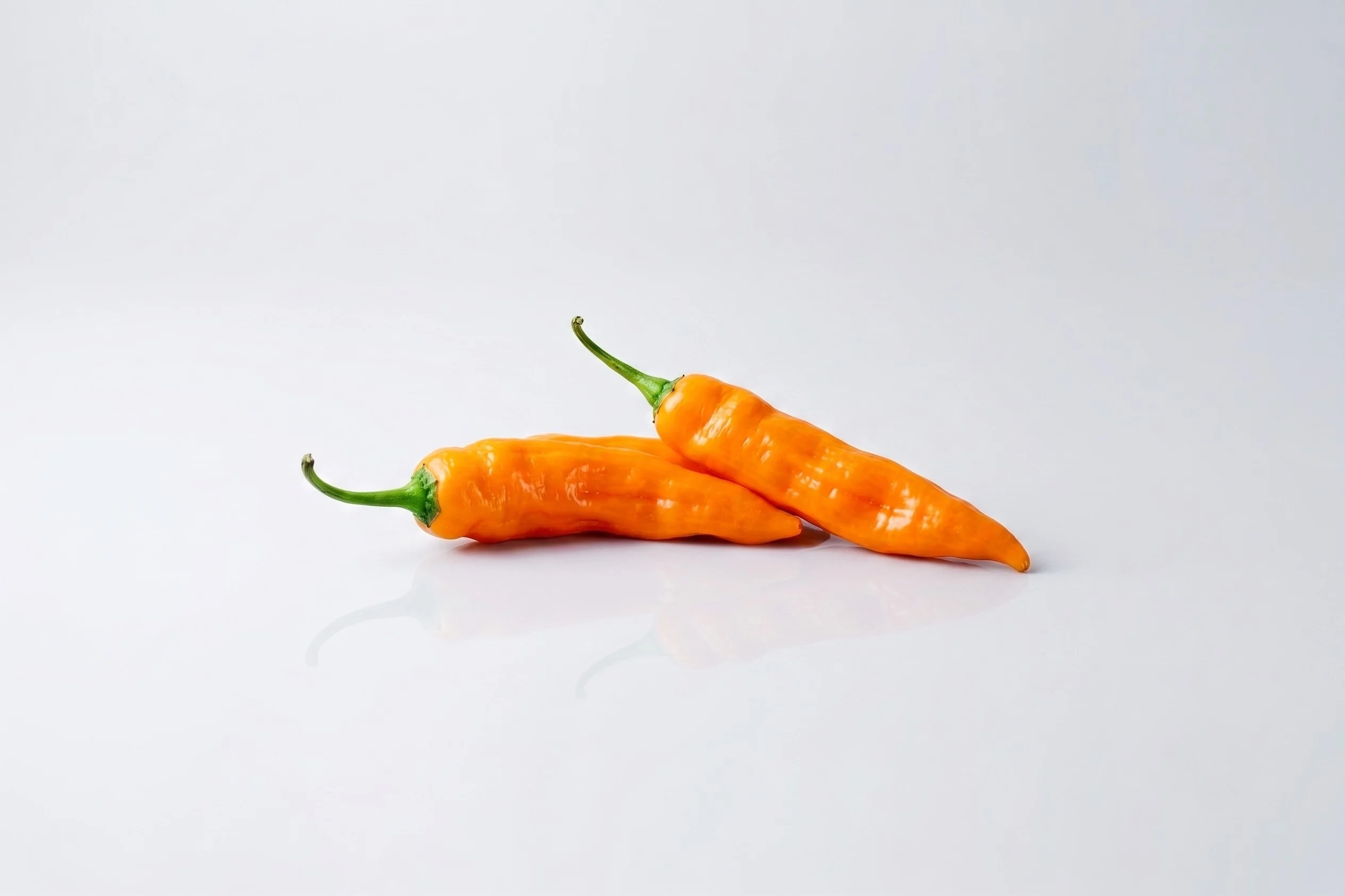 Two bright orange aji amarillo peppers with green stems on a white reflective surface against a light grey background.