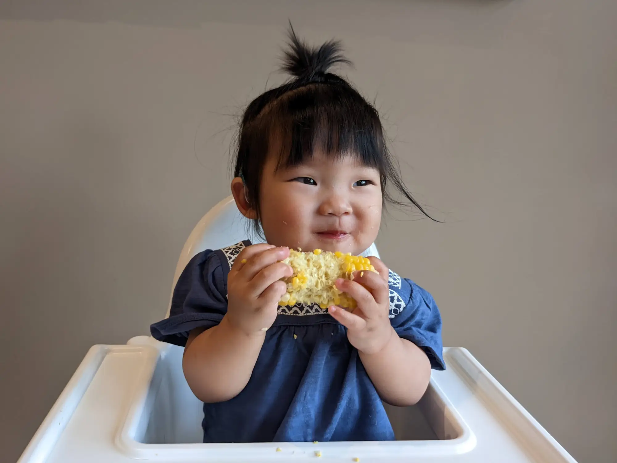 Smiling baby sitting in high chair practicing baby led weaning by holding/eating a piece of corn on the cob.