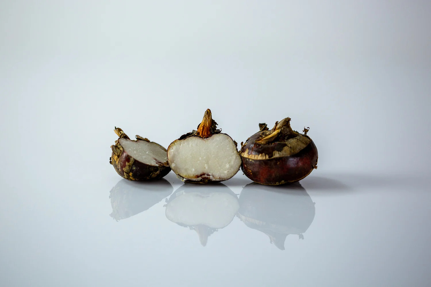 Three water chestnuts on a white reflective surface, with the center one sliced in half to reveal its crisp, white interior, flanked by two whole unpeeled chestnuts with dark brown skin.