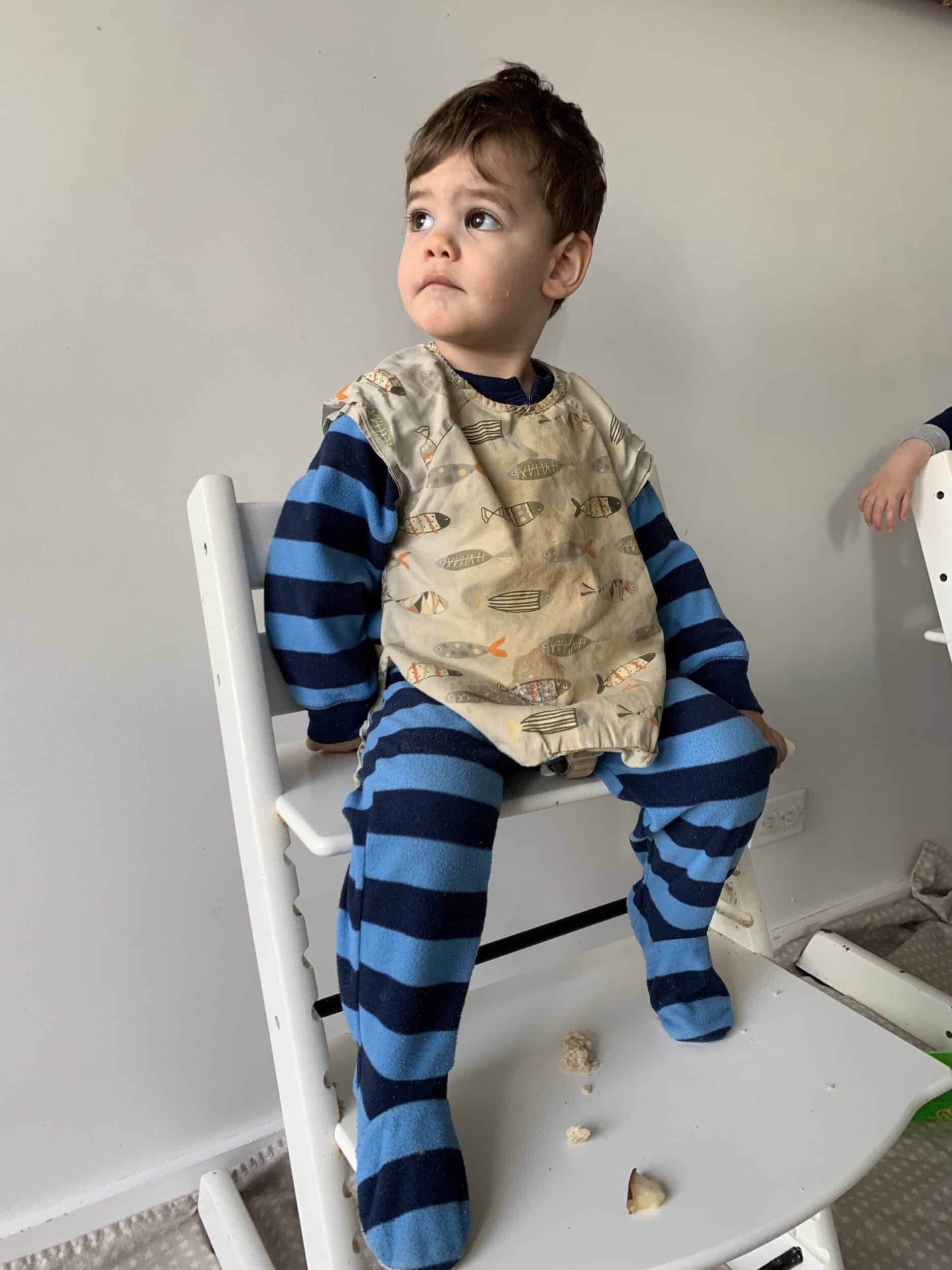 baby in footed pajamas and a bib sitting in a highchair