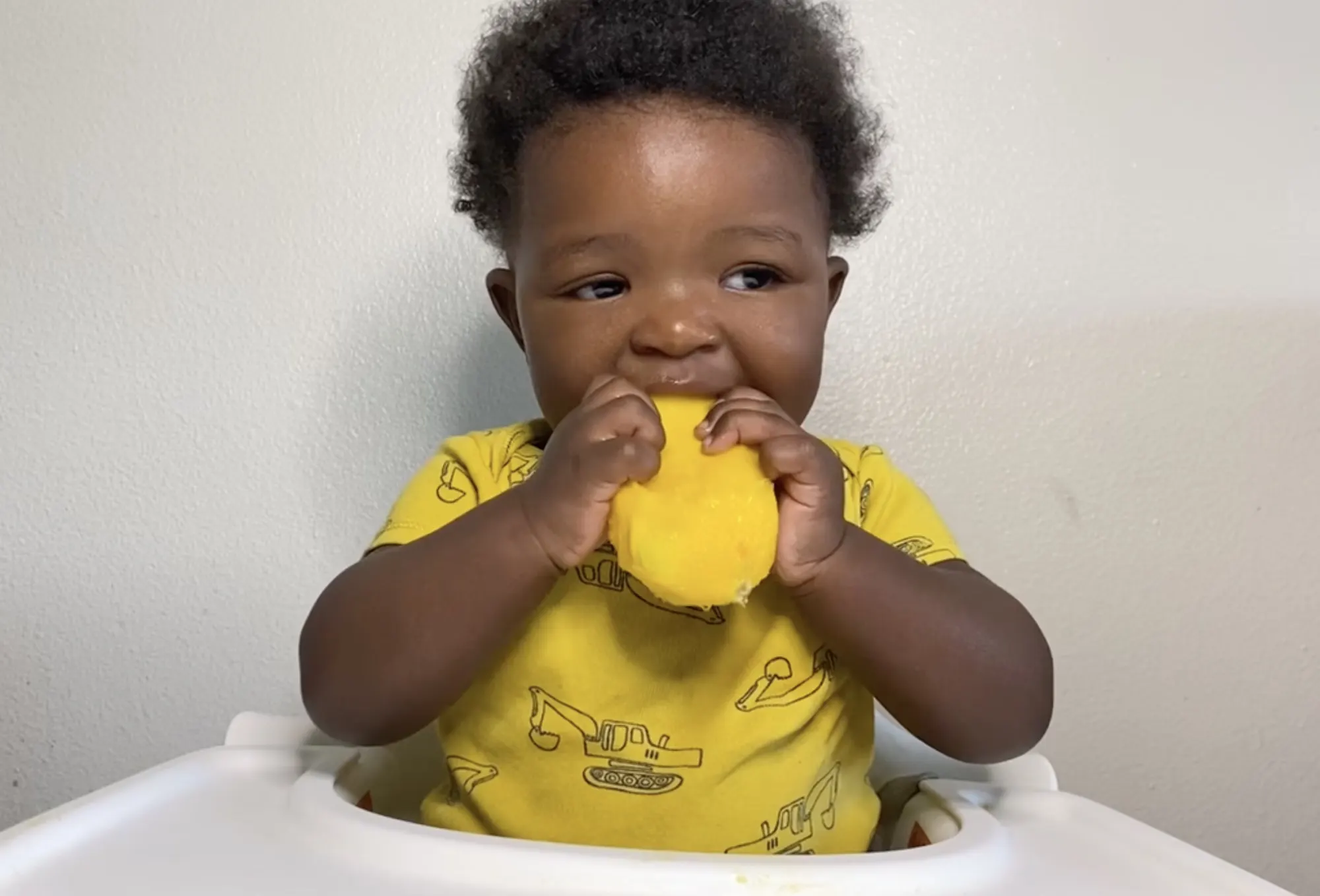 a photograph of a baby in a high chair munching on a mango pit with most of the fruit cut off