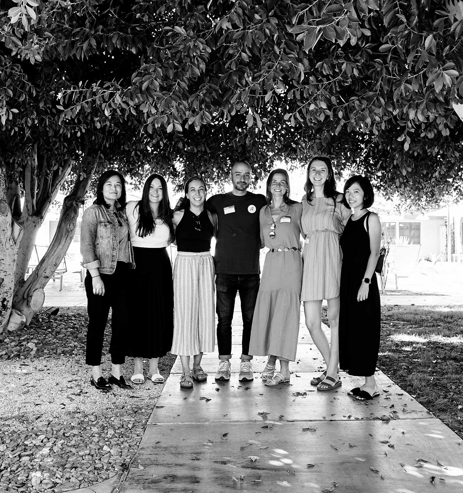 Seven people stand together under the canopy of a tree, smiling at the camera.