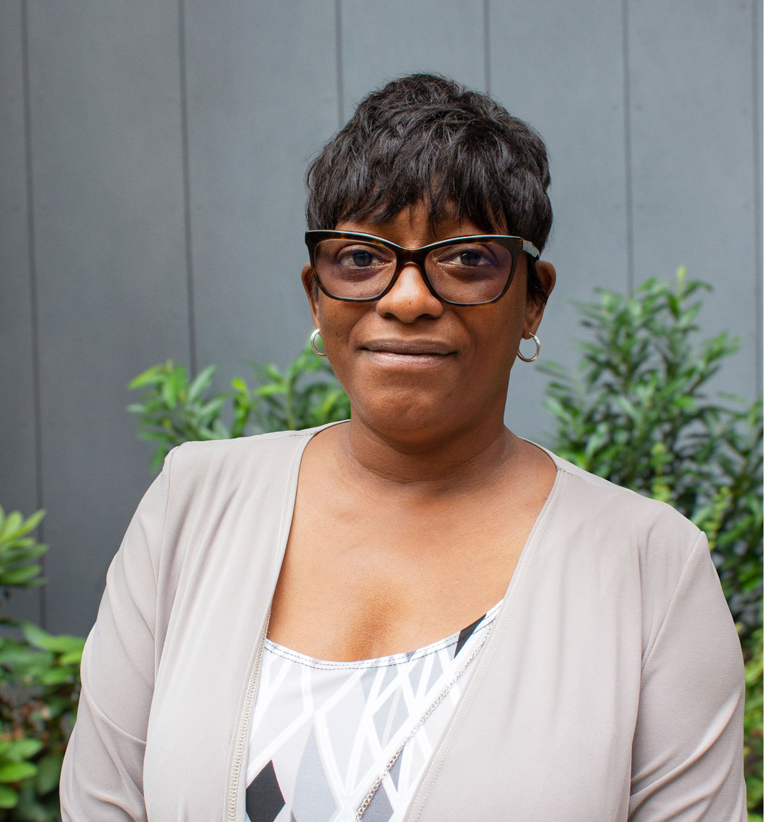 Image of an African-American woman in front of a gray wall and bushes. She is wearing a gray and white shirt with large, black glasses and has short hair.