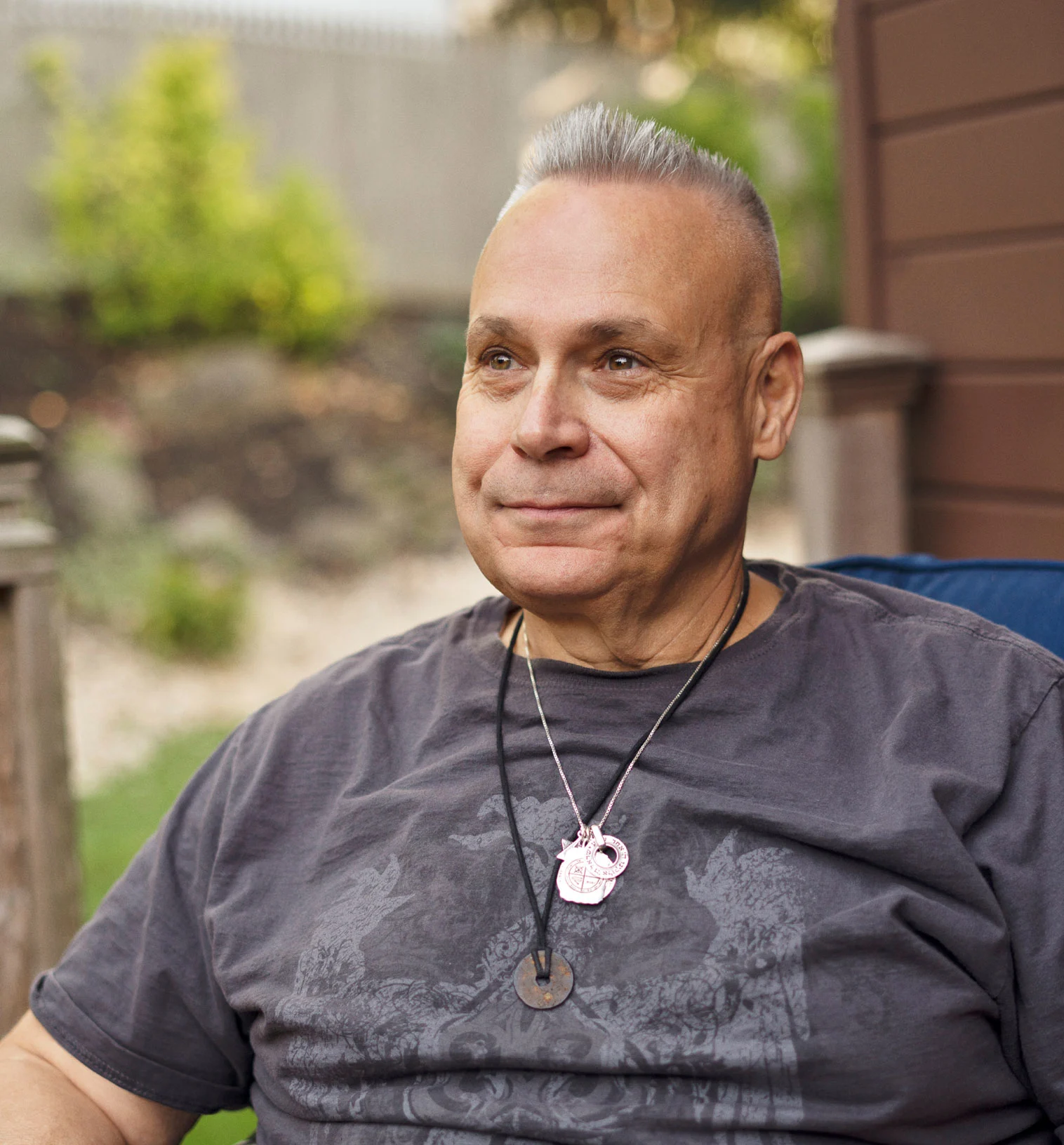 A white man sitting down in a gray shirt with a black rope necklace and a silver necklace.