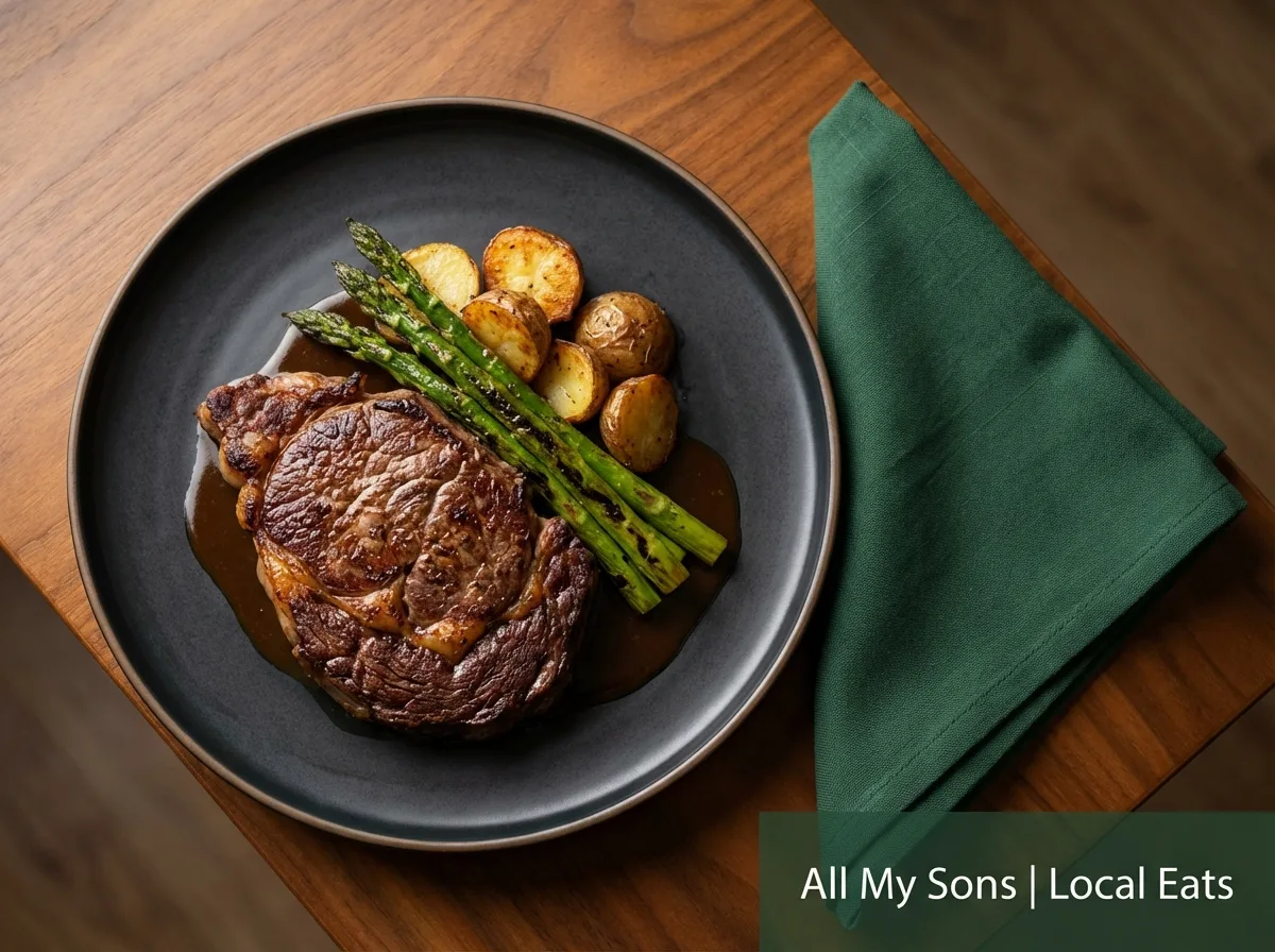 Overhead view of a delicious steak meal on a black plate next to a green napkin, with a banner reading 'All My Sons | Local Eats'.