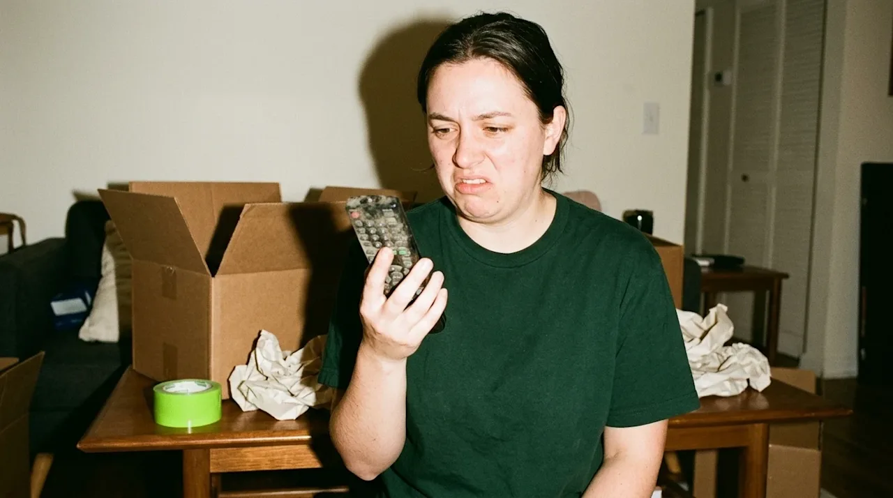 A candid, point-and-shoot style photograph of a person packing for a house move, holding a visibly dusty and grimy TV remote