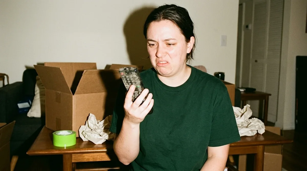 A candid, point-and-shoot style photograph of a person packing for a house move, holding a visibly dusty and grimy TV remote