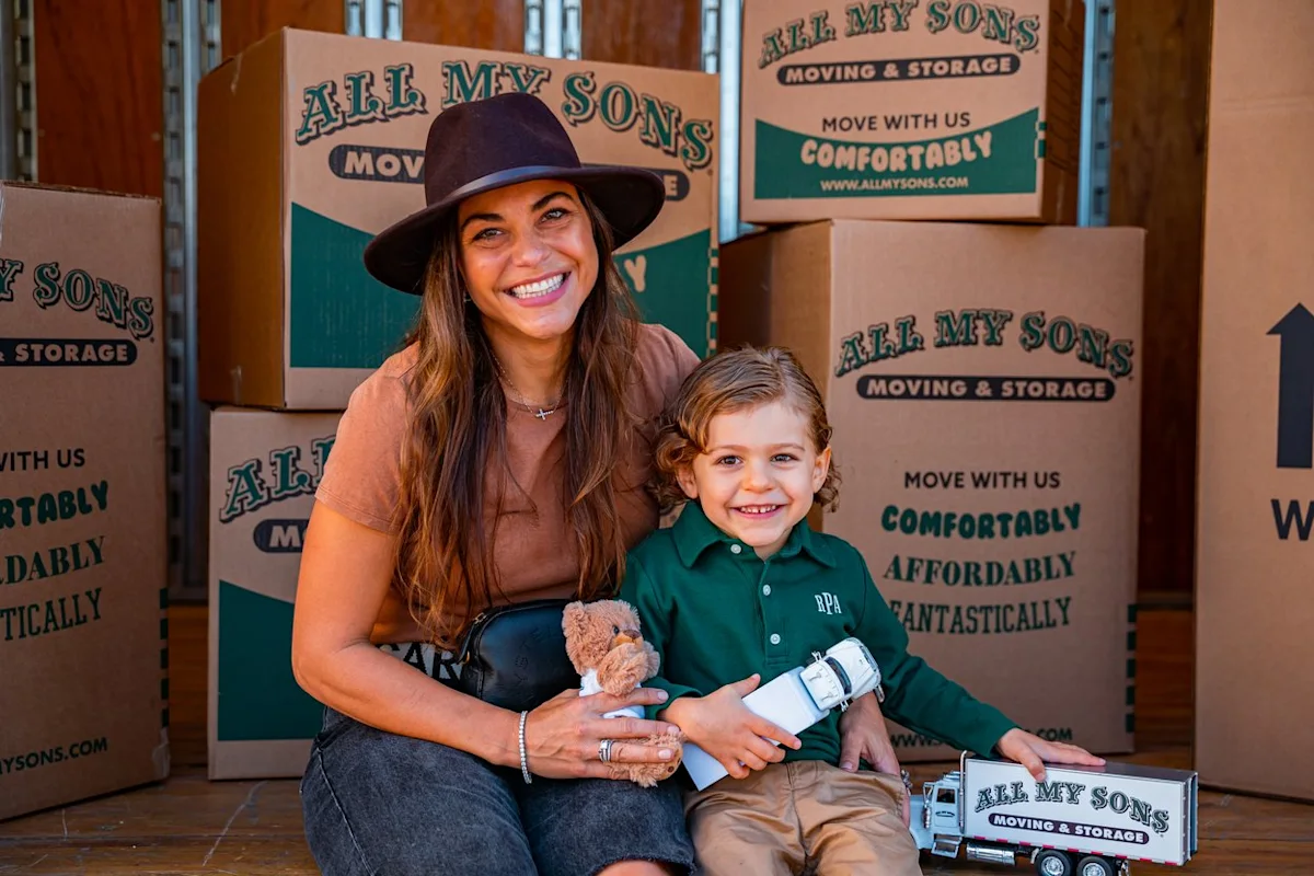 Mother and child sit in the back of an All My Sons moving truck surrounded by moving boxes.