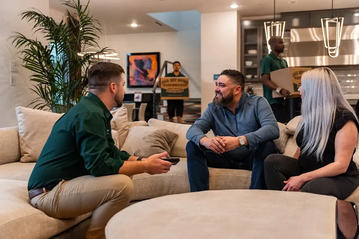 All My Sons team member reviews charges with couple as movers carry boxes into their new house.