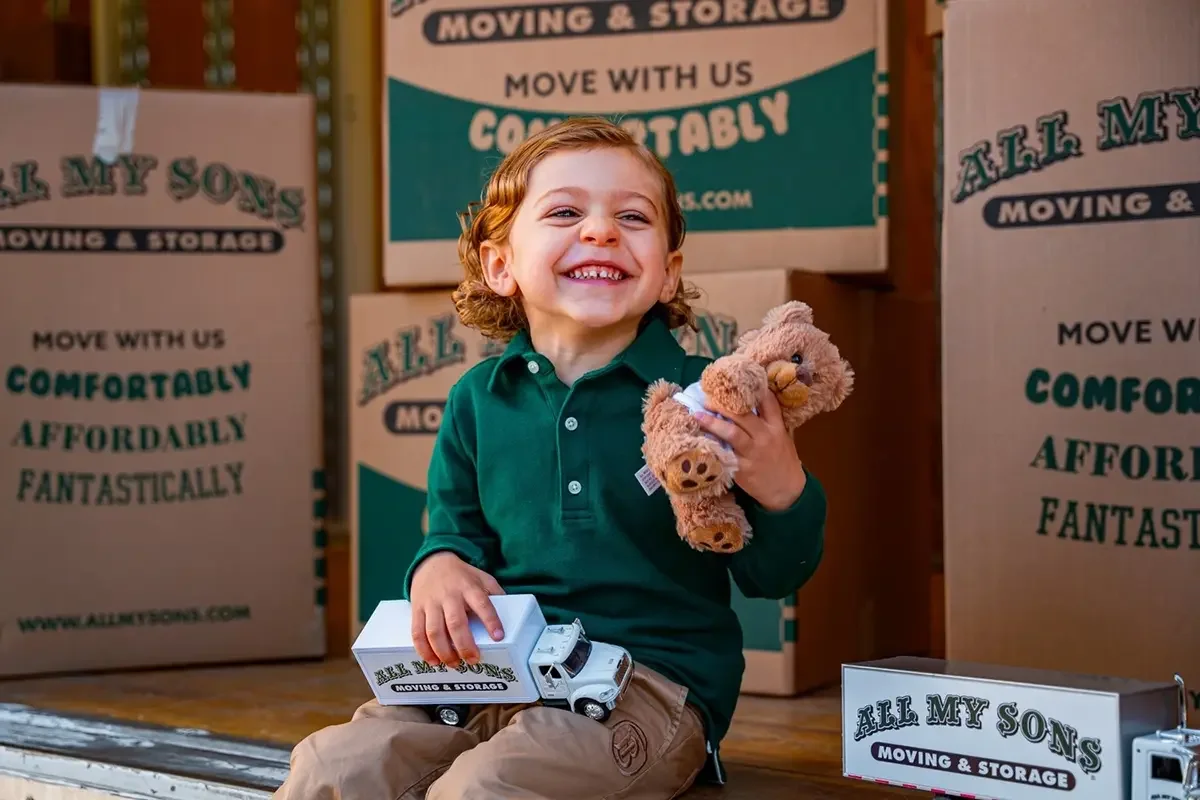 Small boy plays with a toy All My Sons moving truck and teddy bear in the back of a real All My Sons moving truck.
