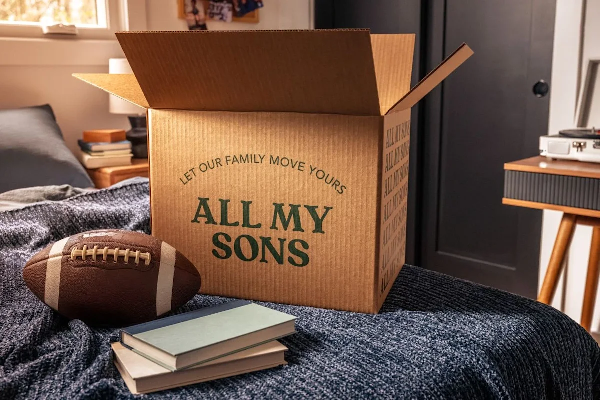 A football and a stack of books await packing in front of an All My Sons Moving and Storage cardboard moving box.
