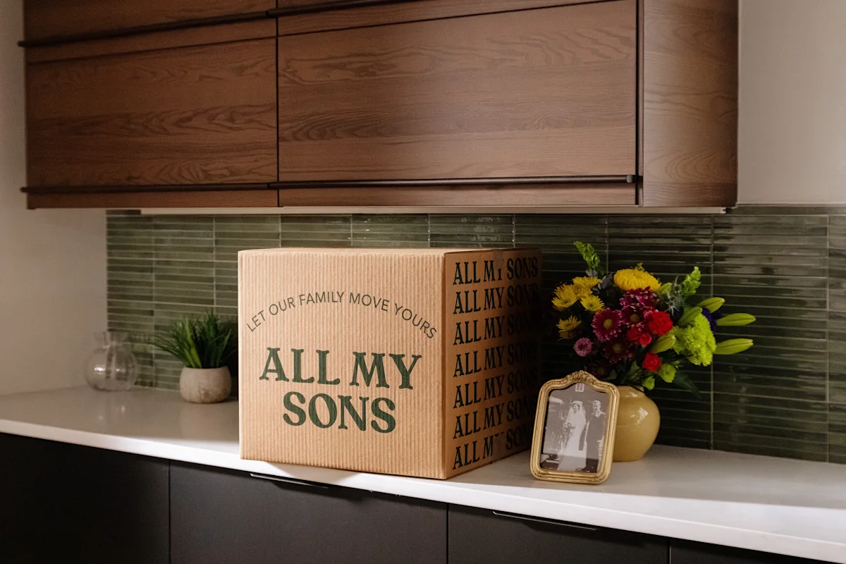 An All My Sons moving box on a kitchen counter next to a vase of flowers and a wedding picture.