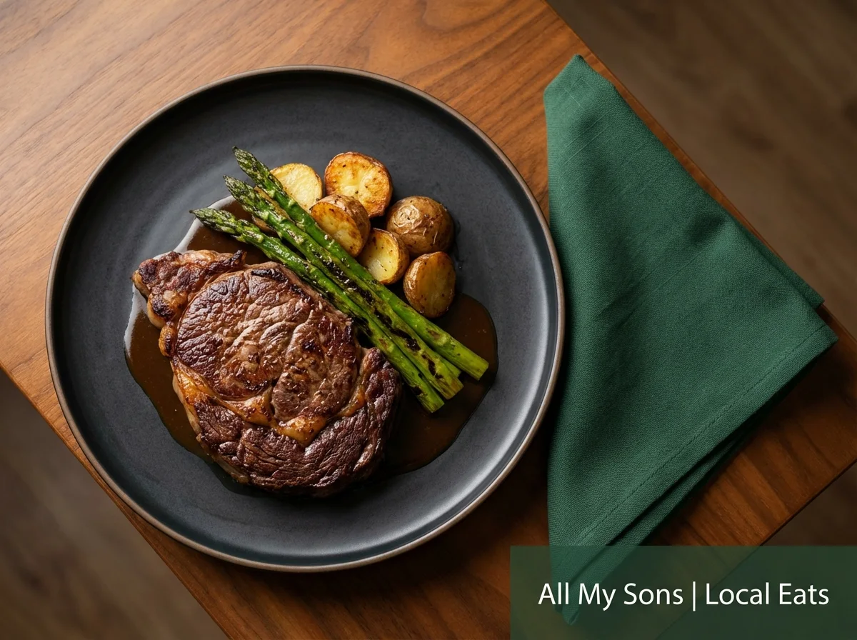 Overhead view of a delicious steak meal on a black plate next to a green napkin, with a banner reading 'All My Sons | Local Eats'.