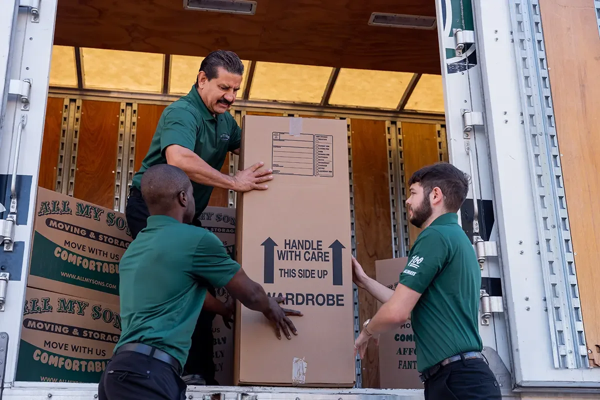 Professional movers from All My Sons Moving and Storage pack a wardrobe box into the back of a moving truck.