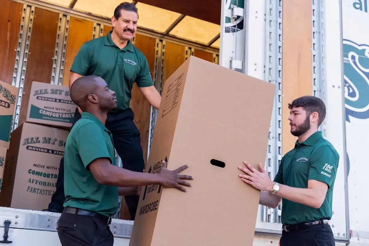 Three All My Sons movers load a wardrobe box into the back of a moving truck.