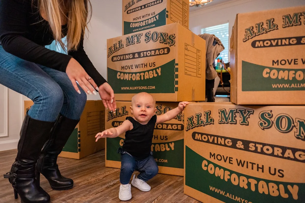Woman with rings on helps toddler stand by All My Sons moving boxes.