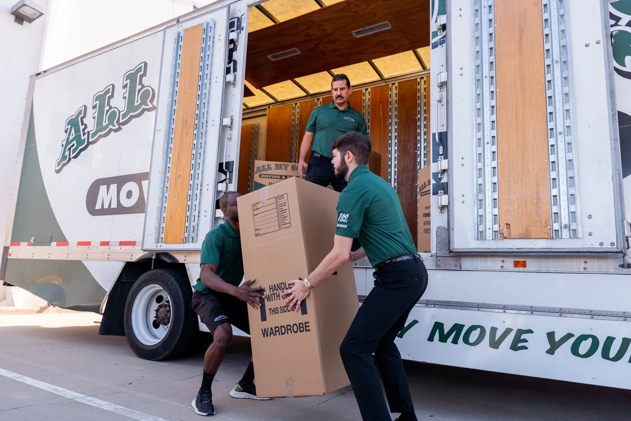 Professional movers from All My Sons Moving and Storage lift a wardrobe box of clothes into a moving truck.