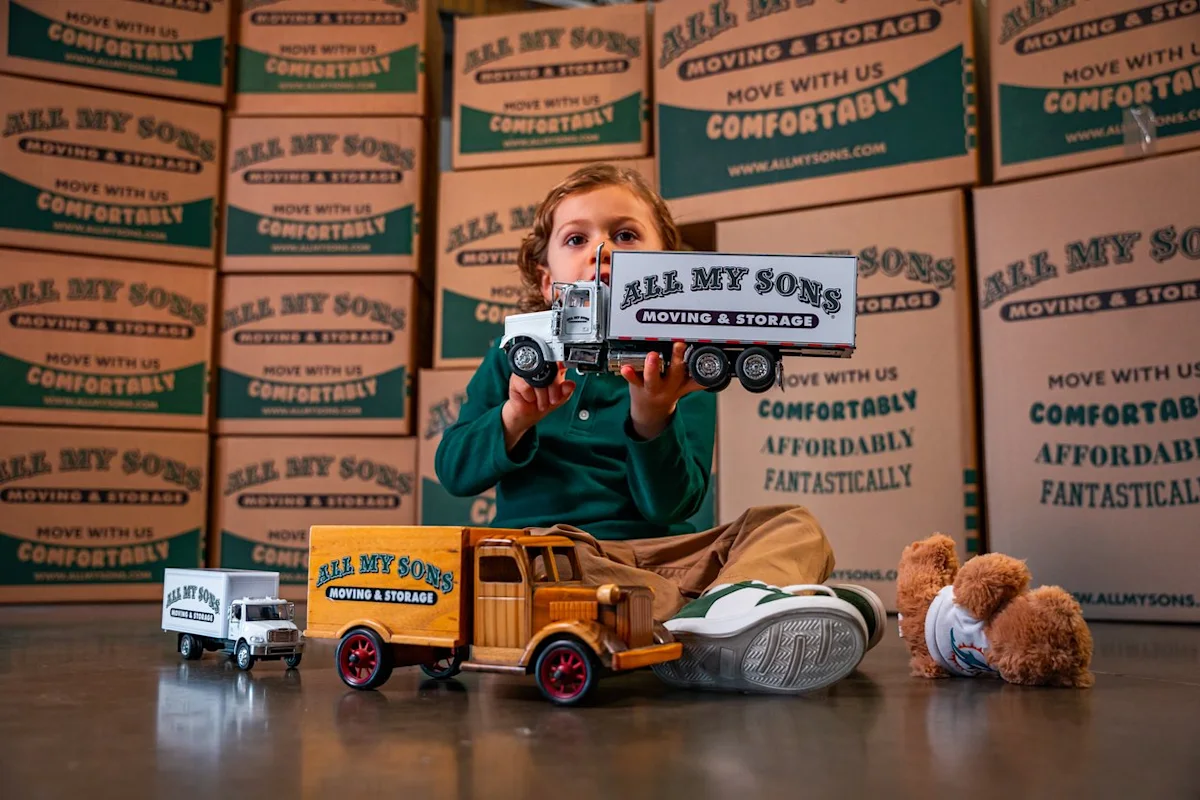 Small child plays with tiny, dollhouse-sized trucks in front of a wall of moving boxes from All My Sons Moving and Storage. 