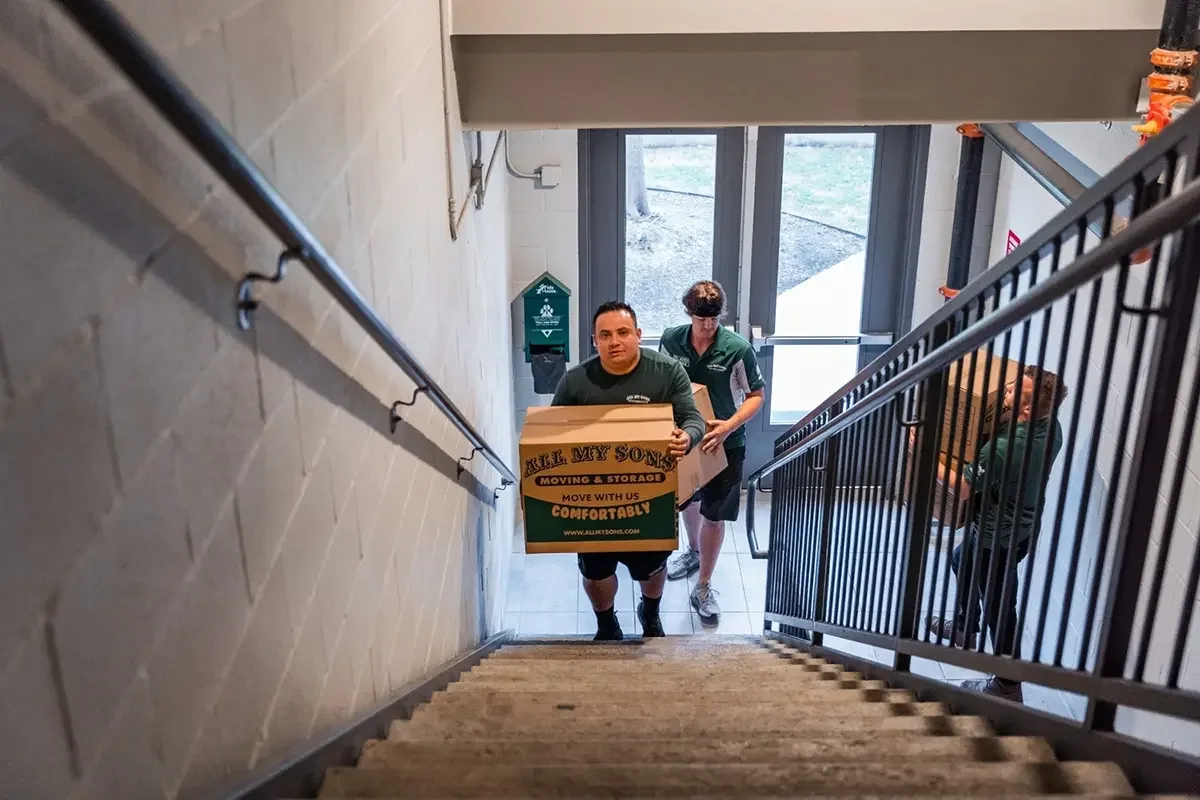 All My Sons mover carries a moving box up a long flight of stairs in an apartment complex.