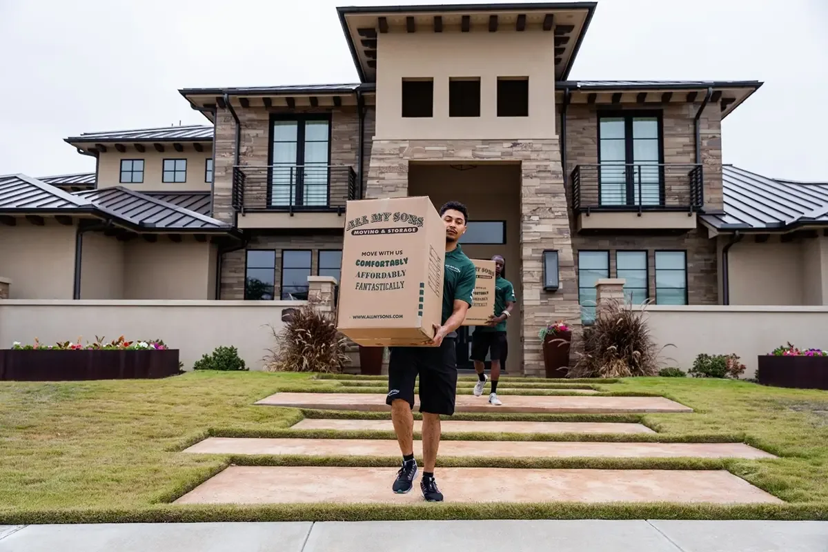 All My Sons professional movers carry large boxes out of a brown-bricked house.