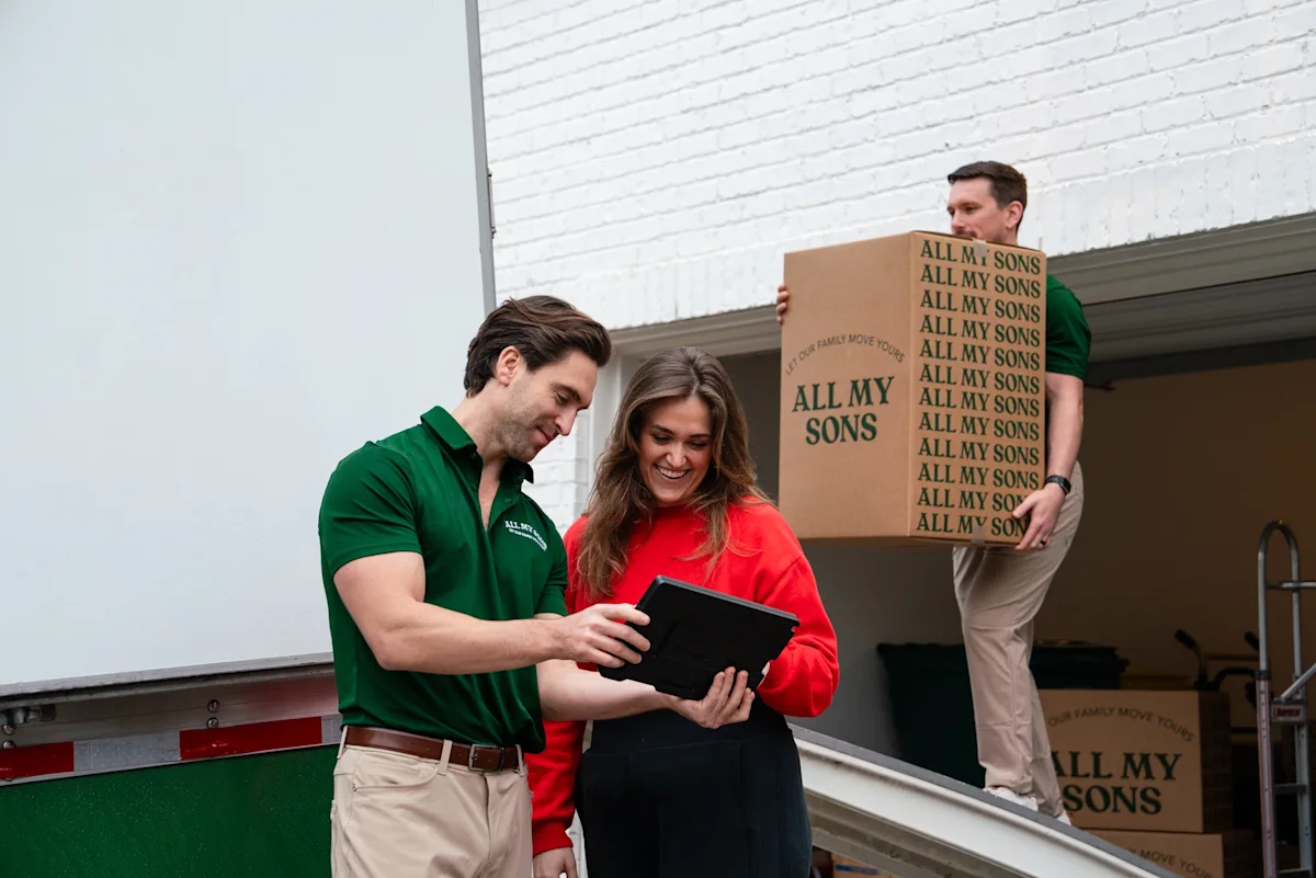 An All My Sons movers explains terms and pricing to a customer while another mover loads boxes in the truck.