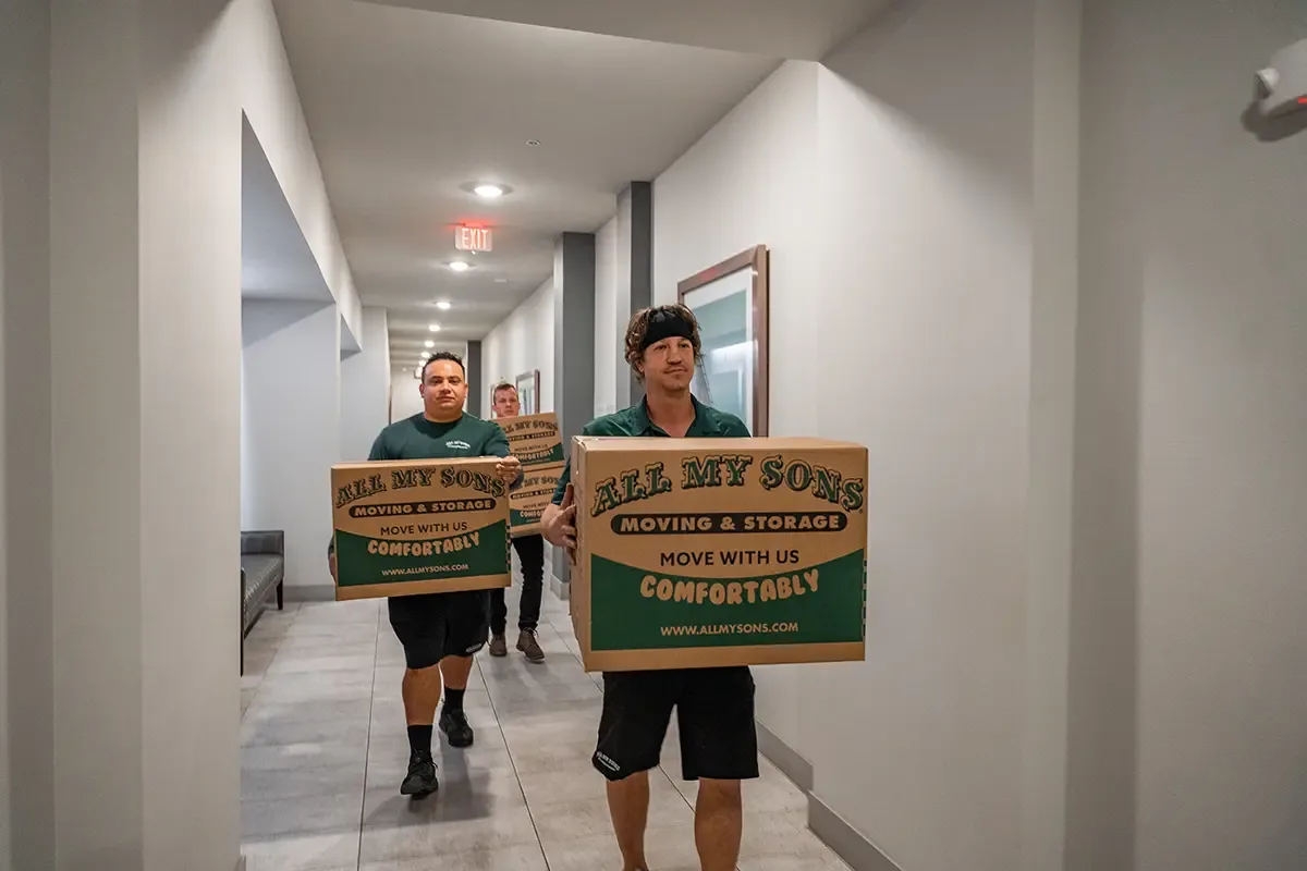 Professional movers from All My Sons carry boxes of moving essentials down a hallway.
