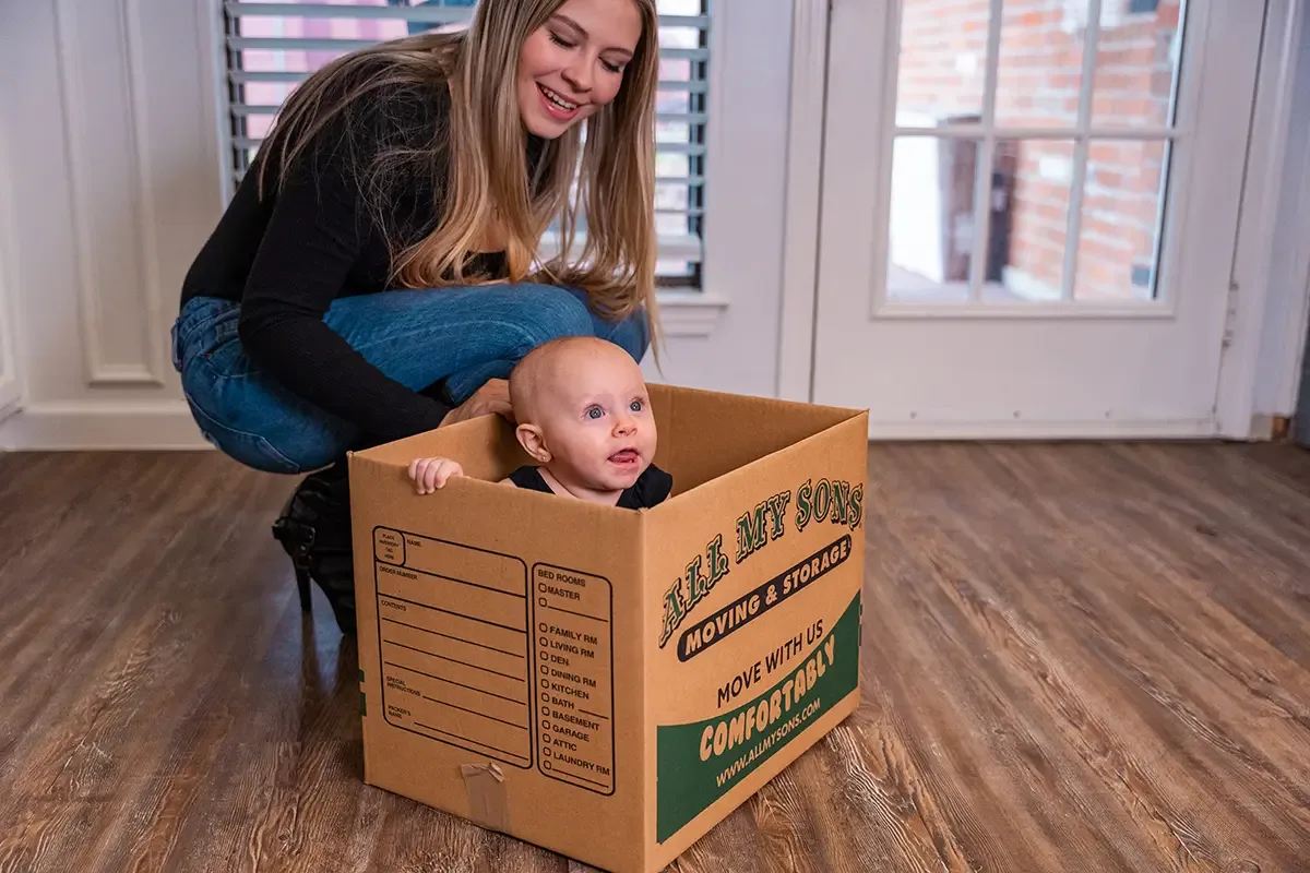 A toddler peeks out of an All My Sons moving box while their mother watches.
