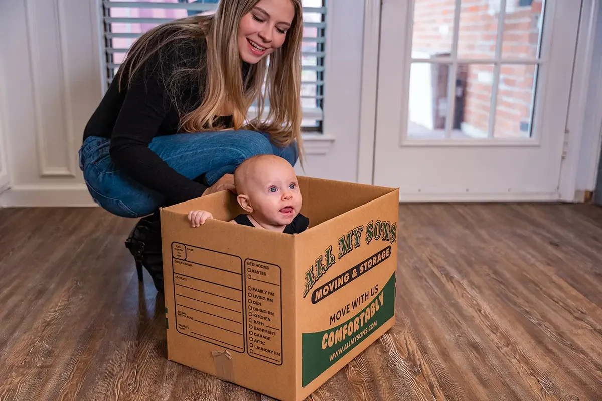 A toddler peeks out of an All My Sons moving box while their mother watches.