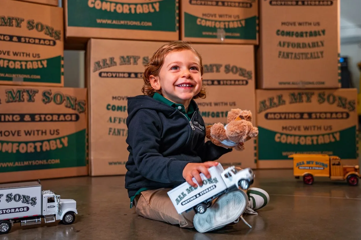 Child plays with a toy All My Sons Moving and Storage truck while surrounded by moving boxes.