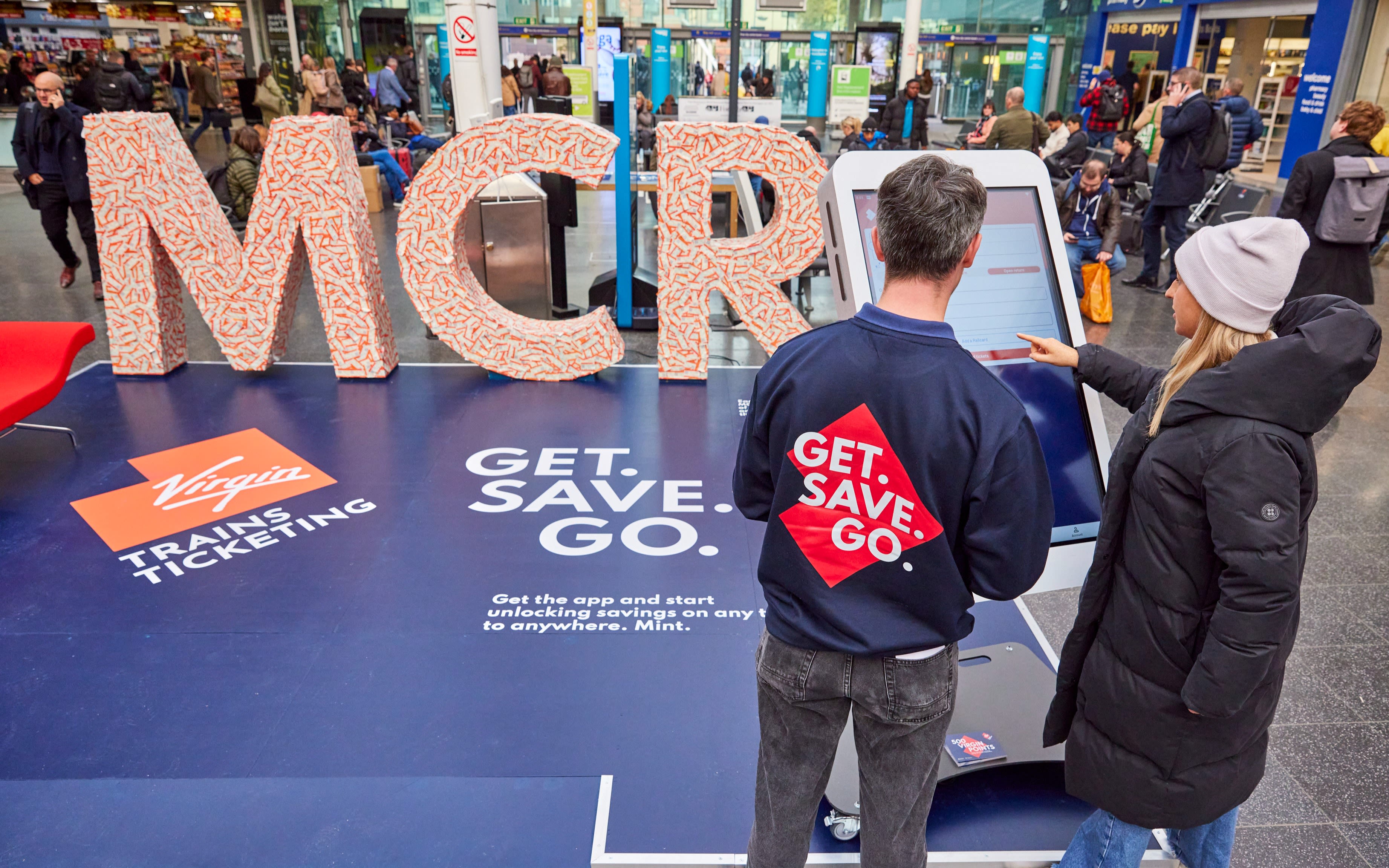 Member of the public and Virgin Trains Ticketing representative standing in front of Virgin Trains Ticketing pop up in Manchester Piccadilly Station