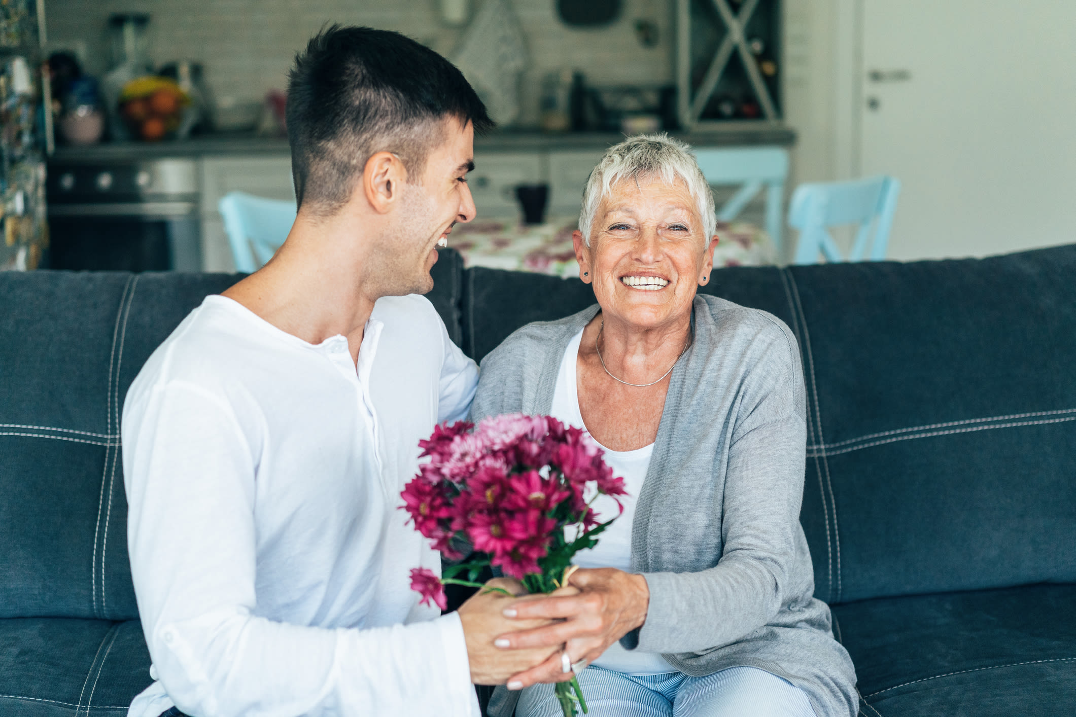 A man sits on a sofa, handing a bouquet of flowers to his mum