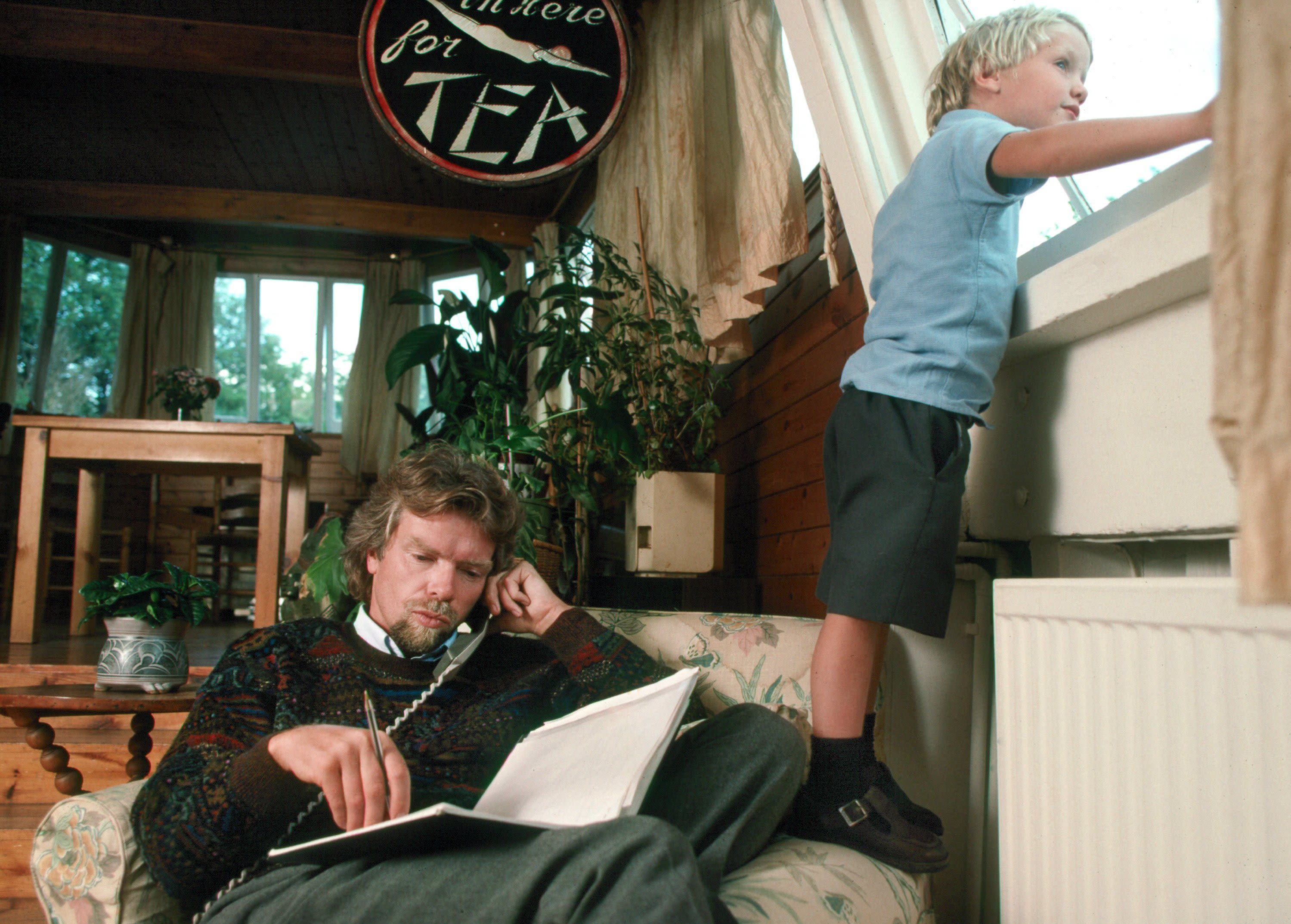 Richard Branson sitting in a chair on the phone in his houseboat. Sam Branson as a boy, is standing on the arm of the chair looking out of the window