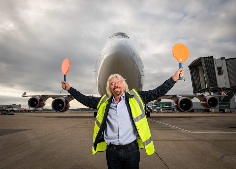 Richard Branson wearing a high visibility jacket holding panels, directing a plane
