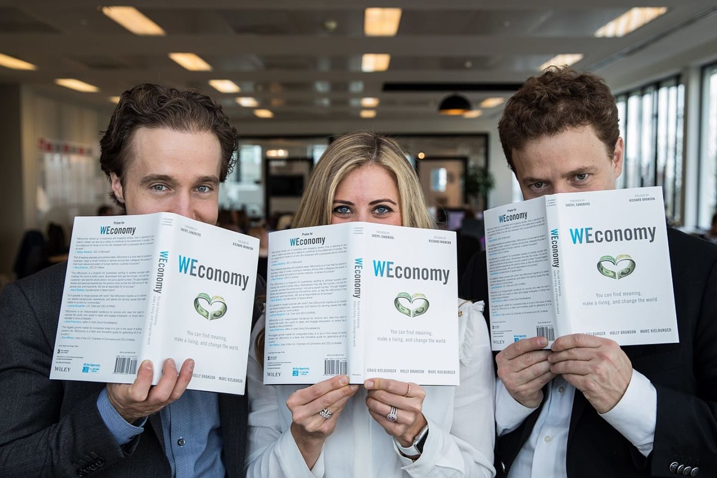 Holly Branson and Marc and Craig Kielburger holding copies of Weconomy in front of their faces, with only their eyes visible