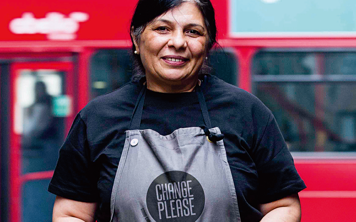 Lady wearing an apron that says change please and standing in front of a red London Bus