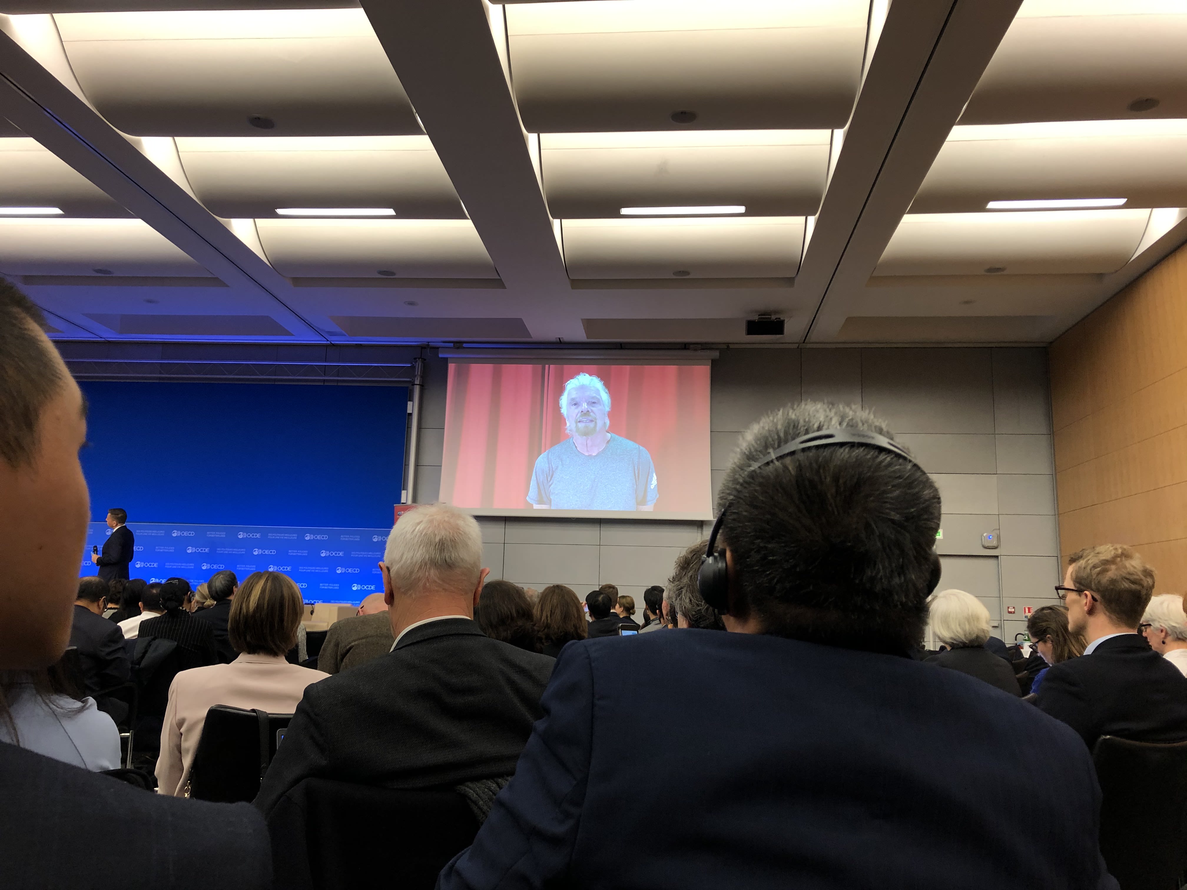 Richard Branson on a video screen at the front of a room. The audience is looking at him, with their backs to the camera