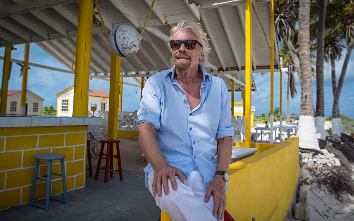 Richard Branson sitting on a yellow wall with the sea in the background