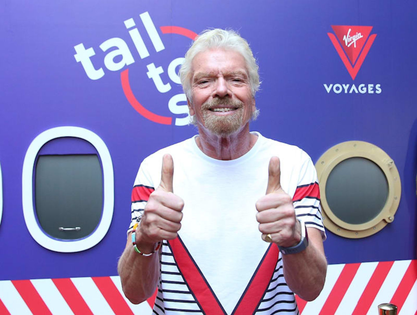 Richard Branson wearing a Virgin Voyages uniform shirt smiling with his thumbs up