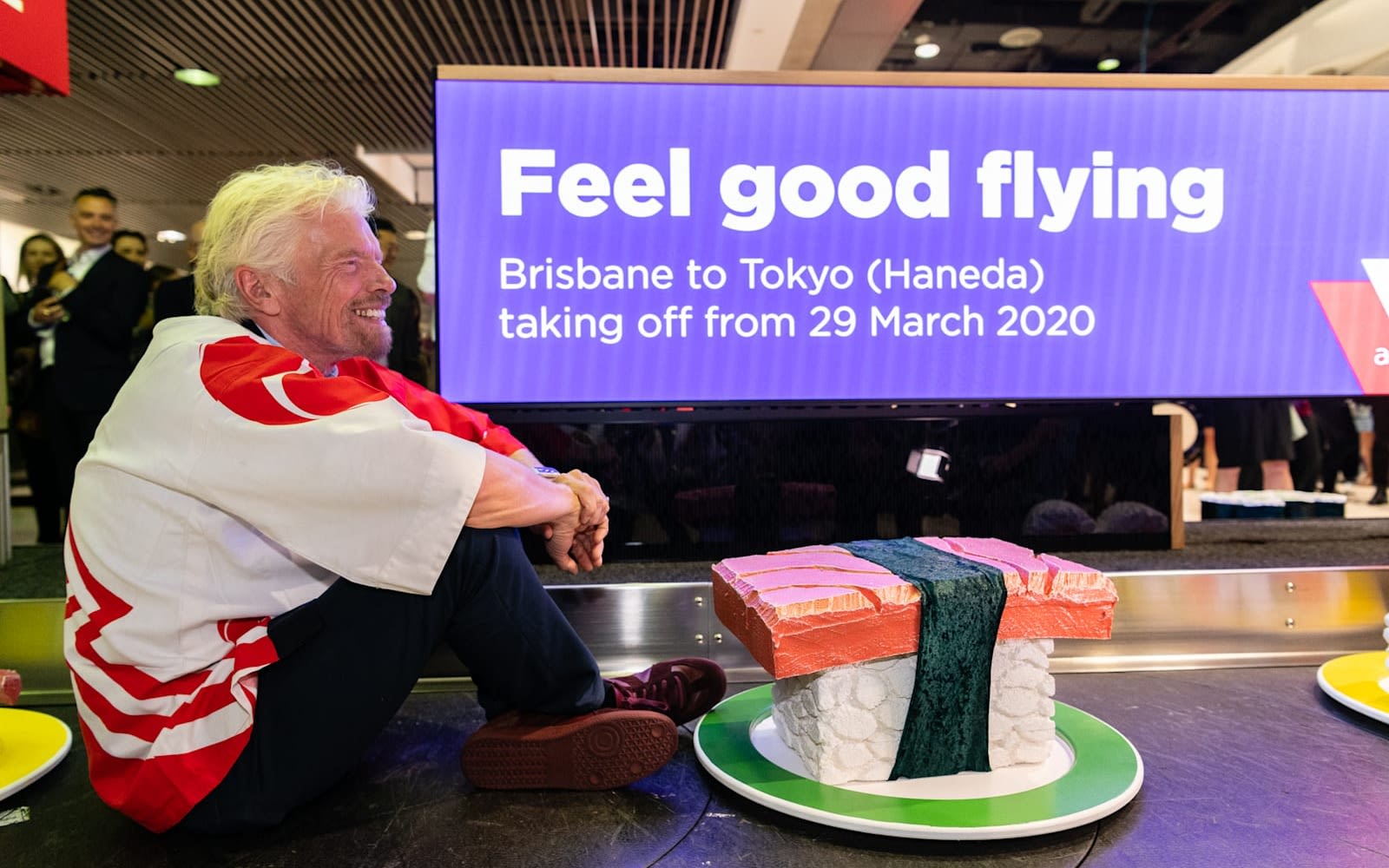 Richard Branson sits on a conveyor belt alongside giant pieces of sushi