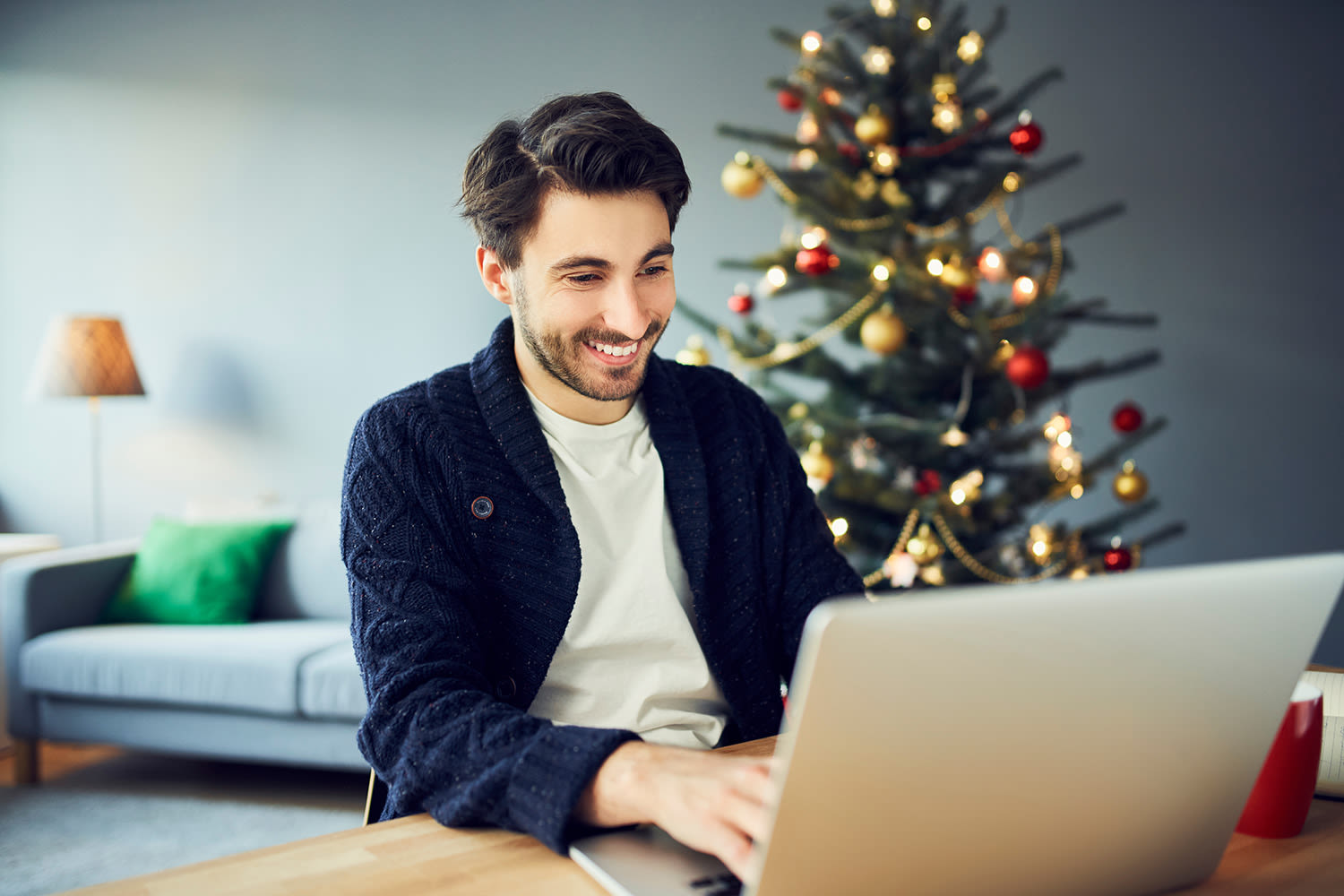 A man using his laptop sitting at a table. A Christmas tree in the background.
