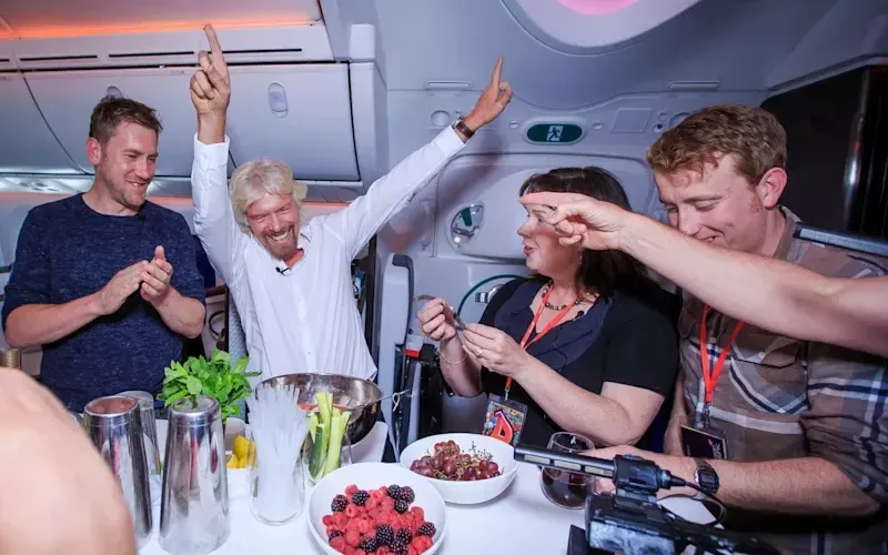 Richard Branson with his hands in the air having fun with other travellers at a bar on a Virgin Atlantic plane