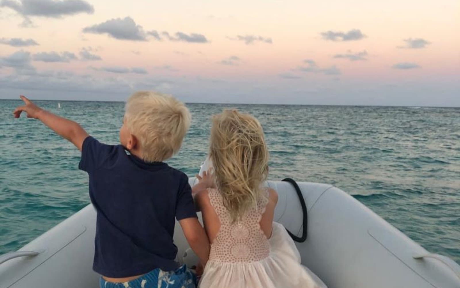 Etta and Artie in a boat with the ocean and horizon in front of them, backs to the camera. Artie is pointing to the left of the image.