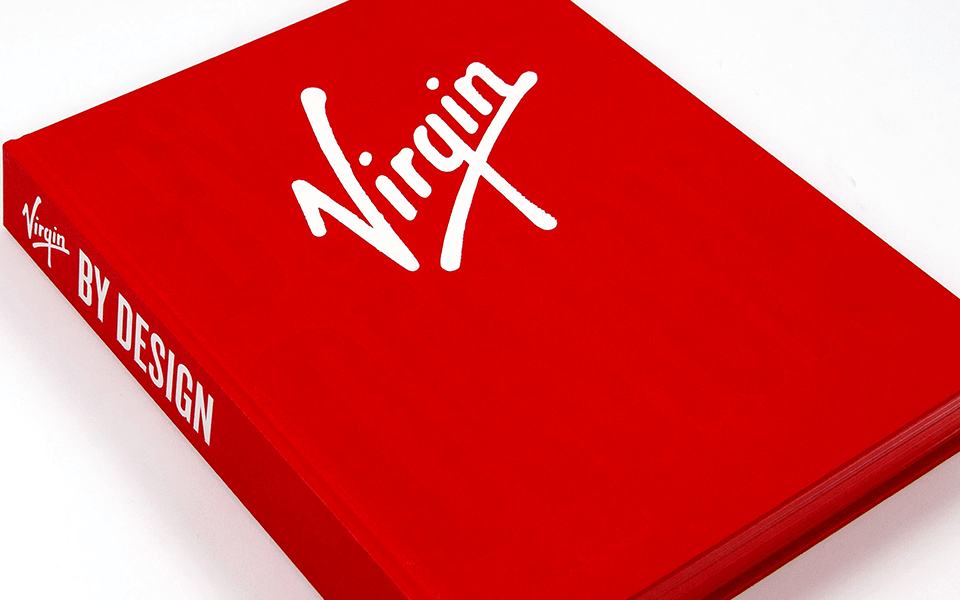 A close up of the Design by Virgin book which has a red cover with the Virgin logo on the front and "Virgin by Design" written on the spine