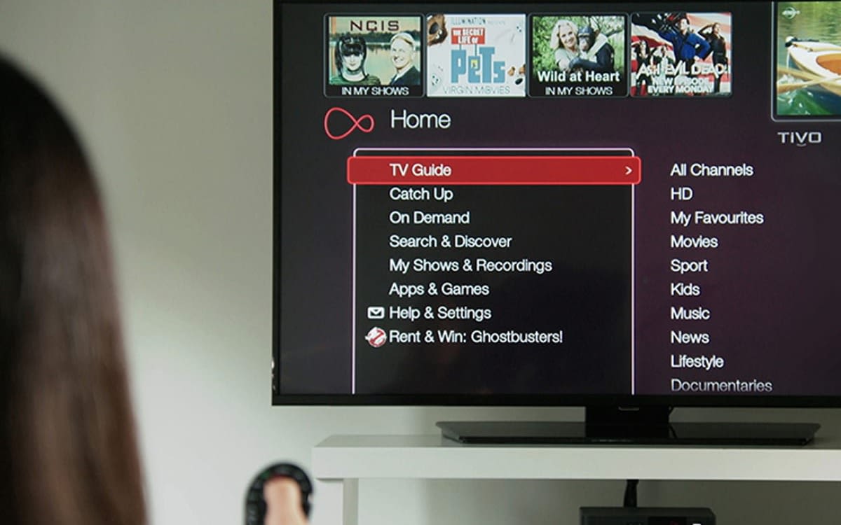 A woman holding a Virgin Media TV remote, the TV shows the Virgin Media TV menu