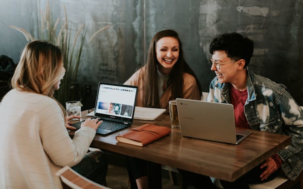 Three women smile together whilst working at their laptops on a shared desk