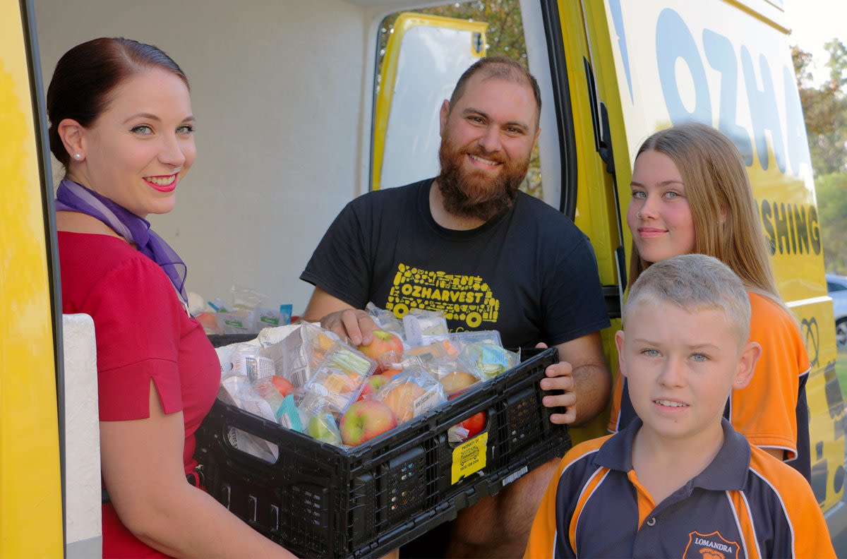 A Virgin Australia cabin crew member hands a basket of food to a man and two children outside a yellow van with the OzHarvest logo